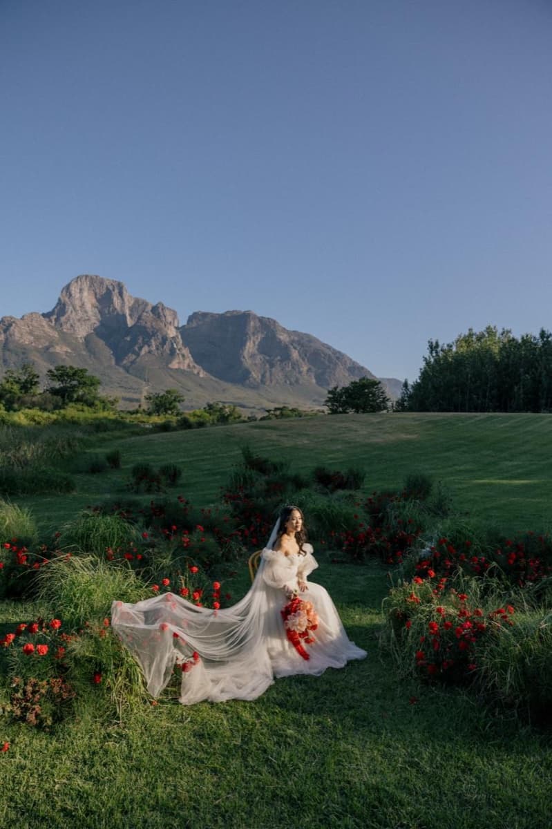 Bride holding a red and pink bouquet in a garden of red flowers beneath mountains